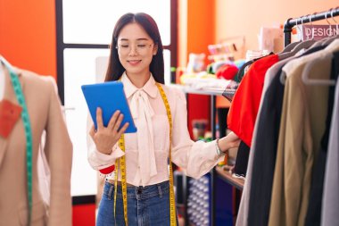 Young chinese woman tailor using touchpad holding clothes on rack at atelier