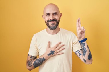 Sarı arka planda dikilen İspanyol dövmeli adam gülümsüyor, elleri göğsünde ve parmakları havada küfür ediyor, sadakat yemini ediyor. 