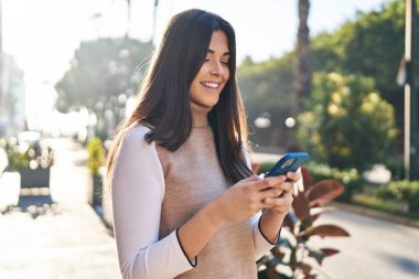 Genç, güzel, İspanyol bir kadın sokakta akıllı telefon kullanarak gülümsüyor.
