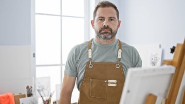 Mature man with grey hair wearing an apron stands confidently in a bright art studio. clipart