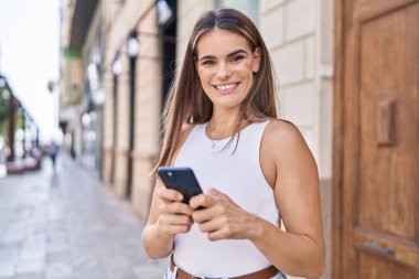 Genç, güzel, İspanyol bir kadın sokakta akıllı telefon kullanarak gülümsüyor.