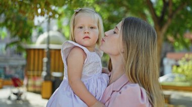 Neşeli anne sarılıyor, kollarında sevgilisiyle dışarıda yeşil parkta kızını öpüyor.
