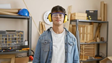 Ciddi yüzlü İspanyol bir genç, yetenekli bir marangoz, gözlük ve kulaklık takıyor, ahşap atölyesinde, kereste ve mobilya inşaat ekipmanlarının arasında rahatça duruyor..