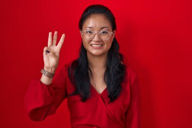 Asyalı genç bir kadın kırmızı arka planda durup üç numarayı işaret ederken kendinden emin ve mutlu bir şekilde gülümsüyor.. 