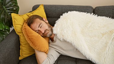 Orta yaşlı bir adam, beyaz bir battaniye ve sarı yastıkla kanepede rahat bir şekilde kestiriyor ve evdeki rahatlığı vurguluyor..