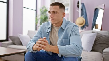 Düşüncelere dalmış genç bir İspanyol adam iyi ışıklandırılmış, modern bir oturma odasında gündelik bir koltukta oturuyor..