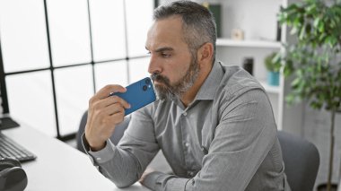 Gri sakallı, düşünceli, olgun bir adam ofisinde akıllı telefon kullanıyor.