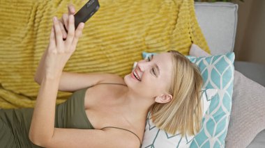 Evdeki kanepede uzanmış akıllı telefon kullanan genç sarışın kadın.