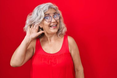 Kırmızı arka planda gri saçlı orta yaşlı bir kadın. Kulağı kulağında bir söylenti ya da dedikoduyu dinliyor. Sağırlık kavramı. 