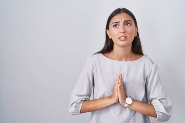 Young hispanic woman standing over white background begging and praying with hands together with hope expression on face very emotional and worried. begging. 