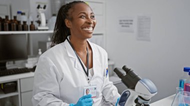 Laboratuvar önlüğü giymiş neşeli bir bilim kadını mikroskop ve ekipmanlarıyla modern bir laboratuvarda araştırma yapıyor..