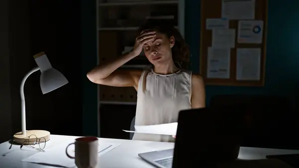 A stressed young woman works late at the office, illuminated by a desk lamp with a laptop, paperwork, and coffee mug.