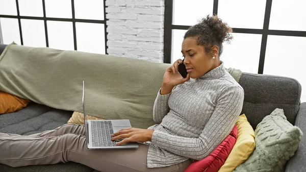 A relaxed woman multitasks with a laptop and phone on a cozy couch indoors.
