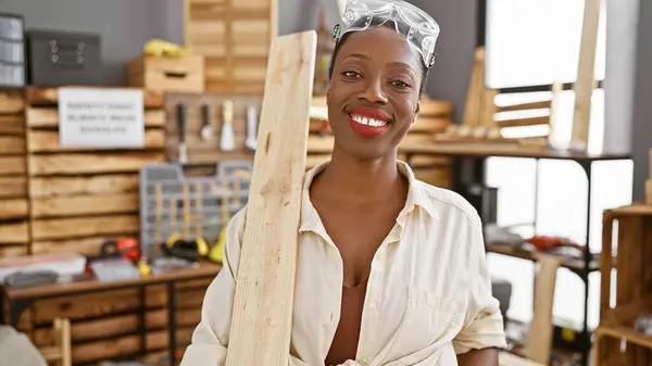 Smiling african american woman carpenter enjoying her job holding wood plank at carpentry workshop
