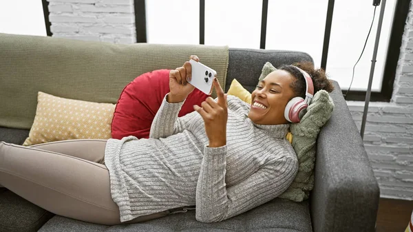 Smiling woman wearing headphones using smartphone on couch indoors.