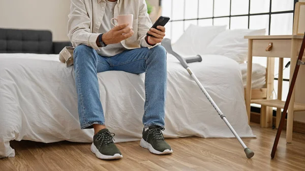 A young man with crutches using a smartphone and holding a mug in a modern bedroom.