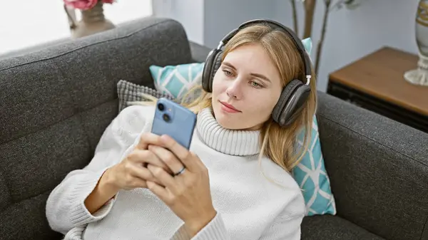Blondýny Žena Sluchátky Používá Smartphone Gauči Zobrazující Ležérní Vnitřní Nastavení — Stock fotografie