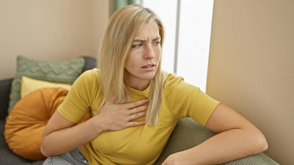 A concerned young caucasian woman experiencing chest pain while sitting at home on a couch.