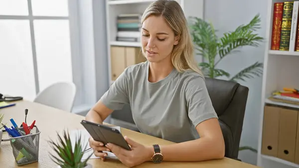 A focused woman using a tablet in a modern office setting with books, plants, and stationary around her.