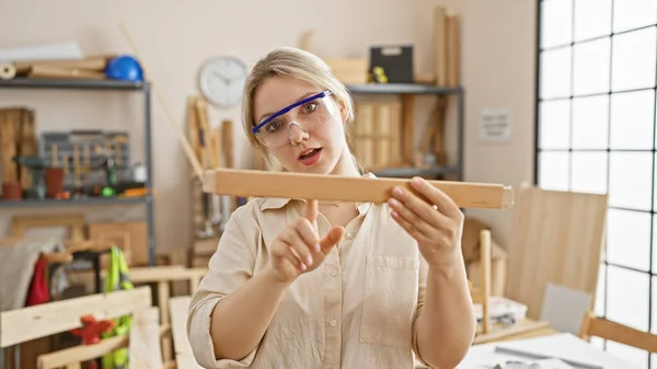 Młoda Biała Kobieta Bada Drewniany Kawałek Jasnym Warsztacie Stolarskim Nosząc — Zdjęcie stockowe