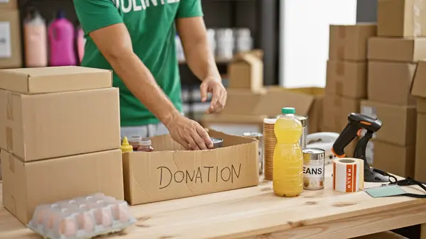 A young man prepares a donation box with food items in a warehouse setting, reflecting charity and community service.