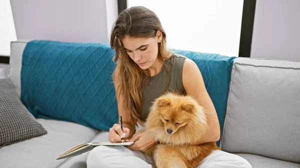 Smart, young hispanic woman, pensively sitting with her dog on the sofa at home, deeply immersed in writing her thoughts in the notebook.