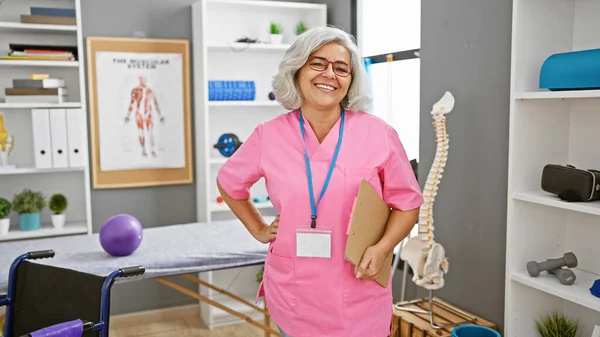 A smiling woman in pink medical scrubs stands confidently in a bright rehabilitation clinic.