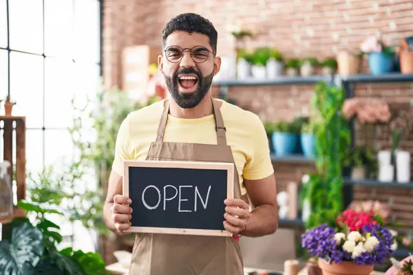 Hispanic man with beard working at florist holding open sign smiling and laughing hard out loud because funny crazy joke.