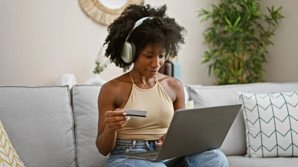 アフリカ系アメリカ人女性が自宅でクレジットカードで買い物をするラップトップとヘッドフォンを使用 — ストック写真