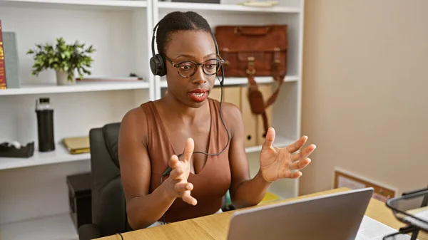 Beautiful african american woman boss wearing glasses, heartily engaged in an online business call. working indoors in her office, she\'s relaxed yet serious, managing her duties via her laptop.