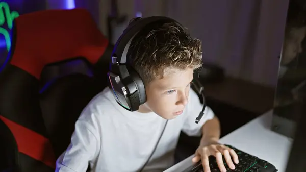 Adorable blond boy streamer, engrossed playing a futuristic video game, streaming live from his digital gaming room at night using tech-gadgets, face focused yet relaxed
