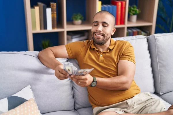 Young latin man counting dollars sitting on sofa at home