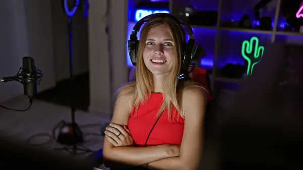 ネオンライト付きダークゲームルームで赤いトップとヘッドフォンを身に着けている金髪の若い女性を笑顔にする — ストック写真