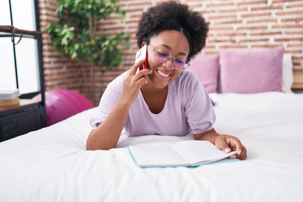 African american woman talking on smartphone reading book at bedroom