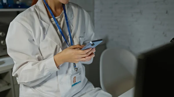 Caucasian woman in lab coat using smartphone indoors at a healthcare facility, displaying professionalism and technology integration.