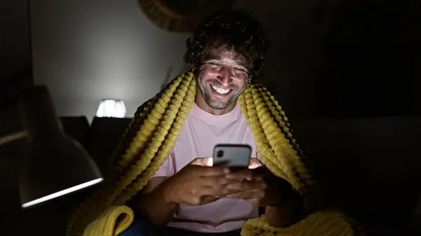 Cheerful Young Hispanic Man Enjoys Browsing His Smartphone Home Wrapped — Stock Photo, Image