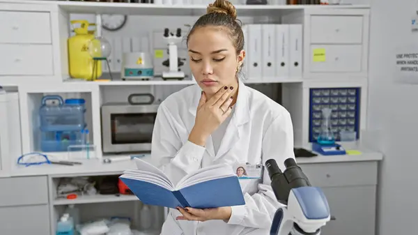 Attractive young hispanic woman scientist, immersed in science research, reading a medical book in a relaxed manner, sitting inside her active laboratories, focused on a promising experiment.