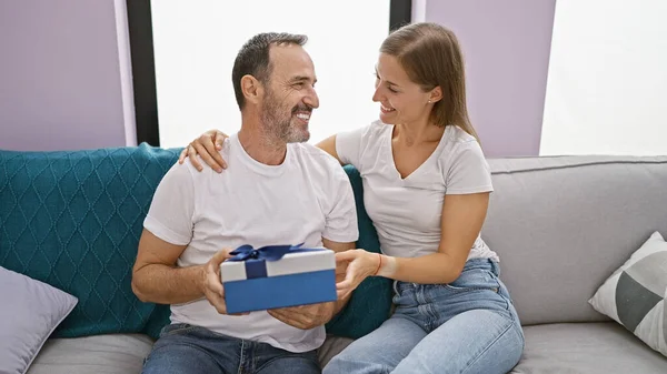 Father\'s loving surprise gift leaves birthday girl, his daughter, smiling and happily surprised on their casual, relaxing living room sofa at home.