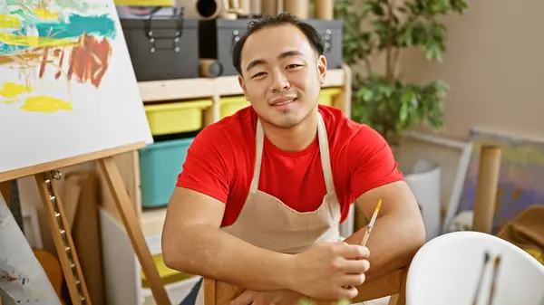Vibrant snapshot, confident young chinese artist, brush in hand, flashing a happy smile, and mastering his craft, sitting in the heart of an indoor art studio