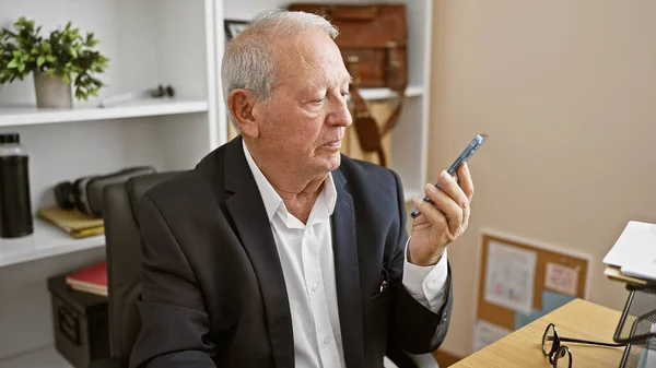 Elegant elder with white hair working hard at the office, serious-faced senior business man sends voice message via smartphone in lively indoor conversation