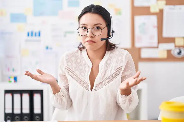 Hispanic young woman working at the office wearing headset and glasses shouting and screaming loud to side with hand on mouth. communication concept.