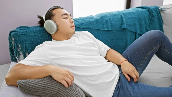 Handsome young chinese man, chilled out while tuned into music on his headphones, sinks into his cozy sofa, at the comfort of his own apartment.