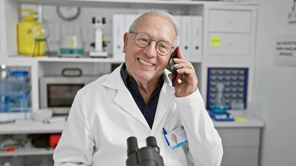 Confident, smiling senior man scientist joyfully engaged in a phone talk, standing in a lab, immersed in profound medical research while smartly working with his trusty smartphone.