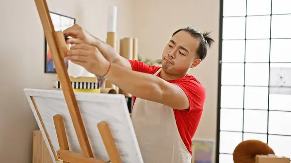 Serious yet relaxed, young chinese artist prepares for a painting session, standing at a studio easel, canvas at hand, concentrating on the unfolding art