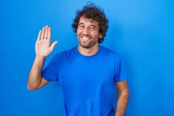 ヒスパニック系の若い男が青の背景に立って手を振っこんにちは幸せと笑顔 優しい歓迎ジェスチャー — ストック写真