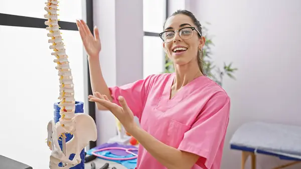 Glowing young hispanic woman physiotherapist explains spinal health with anatomical model at the rehab clinic