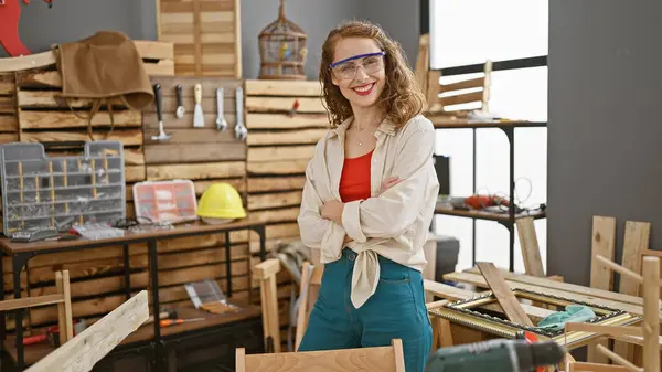 stock image Confident young woman, a professional carpenter, standing relaxed in her woodworking studio, arms crossed, wearing security glasses with a smile