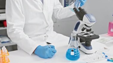 Laboratuvar önlüklü ve eldivenli bilim adamları laboratuarda deney tüpü ve mikroskopla notlar yazıyorlar.