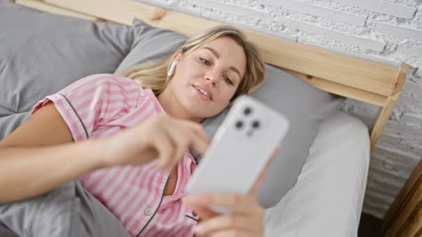自宅でベッドでスマートフォンを使用してパジャマの若いブロンドの女性 リラクゼーションとモダンなライフスタイルを描いた — ストック動画