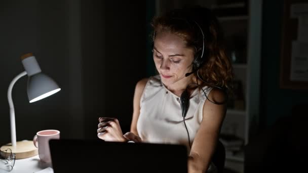 一个带着耳机笑着的年轻女人 晚上在家里的办公室里用笔记本电脑工作 — 图库视频影像